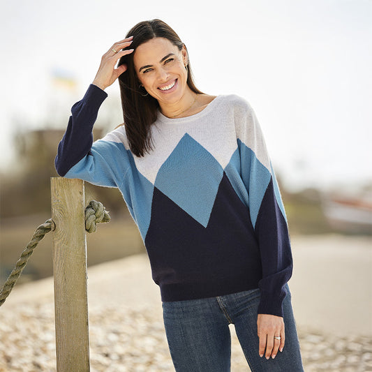 Woman wearing a cashmere diamond blue and navy patterned sweater standing by a wooden post on a beach.