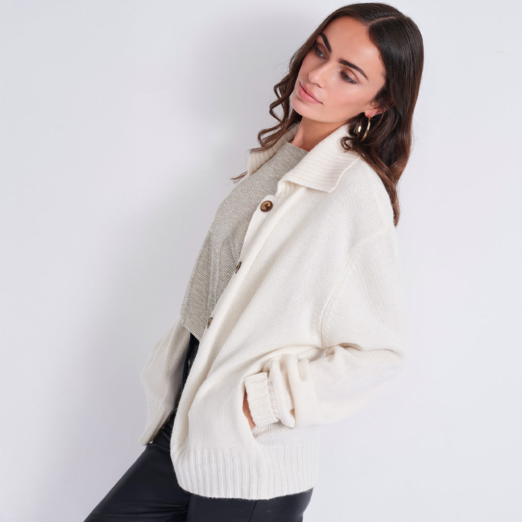 A woman wearing a cream-colored knit cardigan over a gray top and black pants, posing against a light gray studio background.