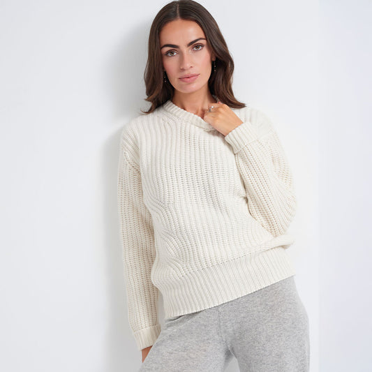 A woman wearing a cream-colored, textured knit crewneck sweater and light gray sweatpants, posing against a plain white wall.