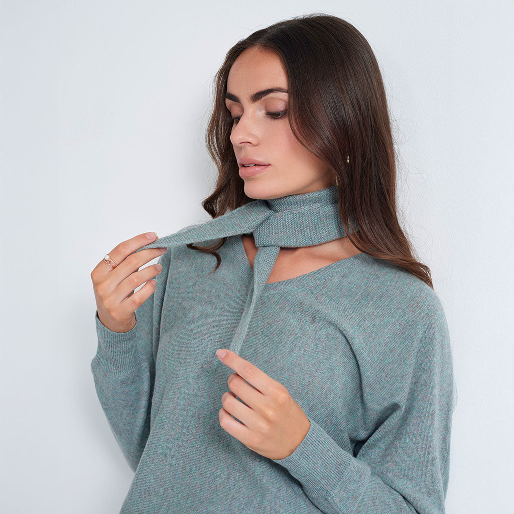 A woman wearing a heathered sage green knit sweater with a high neck and tie detail, posing against a light gray background.