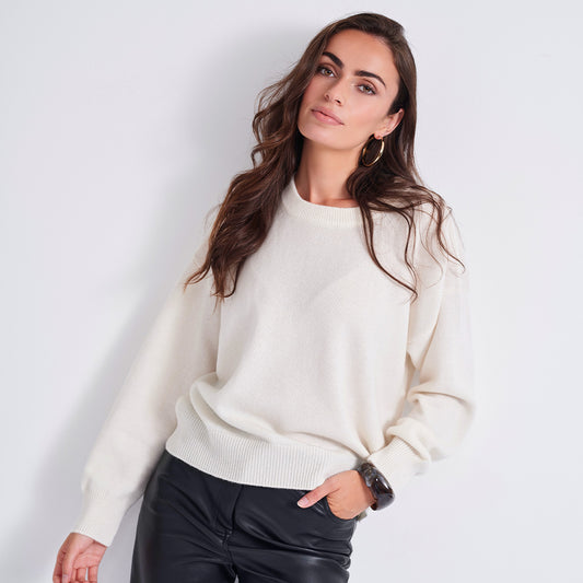 A woman wearing a cream-colored knit crewneck sweater and black leather pants, posing against a light gray studio background.