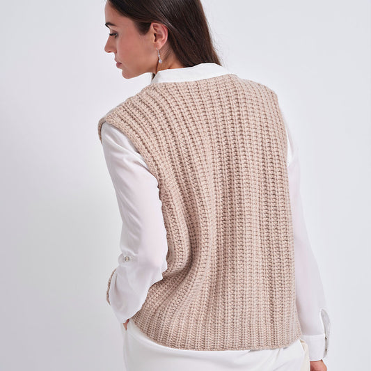A woman shown from the back wearing a beige ribbed knit sleeveless vest over a white collared shirt, against a plain light gray studio background.