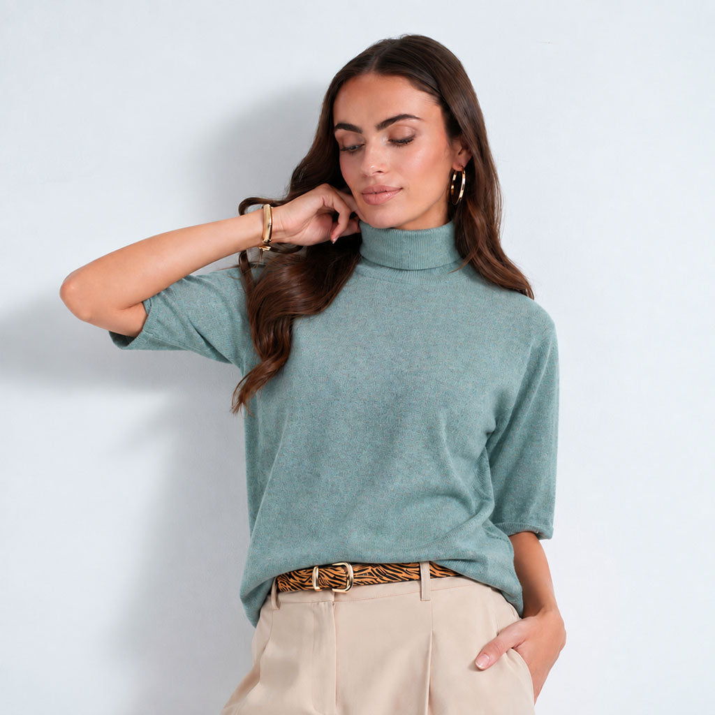 A woman modeling a lagoon short-sleeve mock neck knit top with light beige trousers and a patterned belt, standing against a light blue-gray studio background.