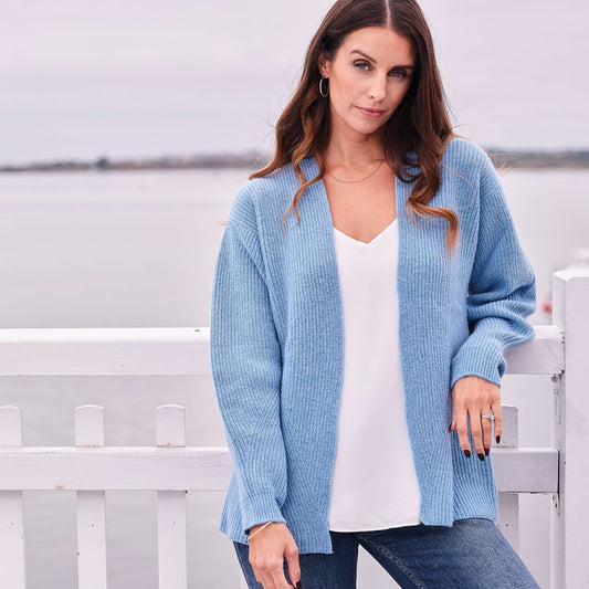 Woman wearing a cashmere light blue cardigan over a white top, standing in front of a white picket fence.