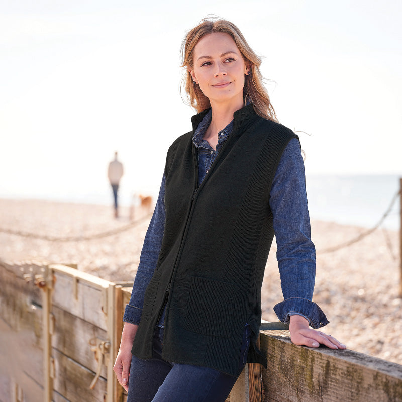 Woman wearing a black zip-up gilet over a blue shirt on a beach.