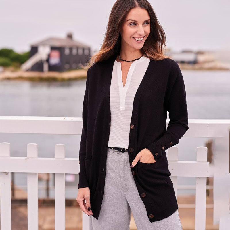 Woman wearing a black cashmere cardigan over a white blouse with gray pants, standing by a white picket fence with water and buildings in the background.