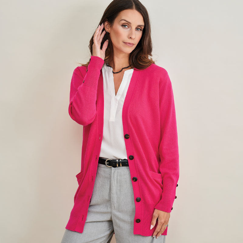 Woman wearing a cashmere bright pink cardigan over a white shirt and gray pants on a beige background