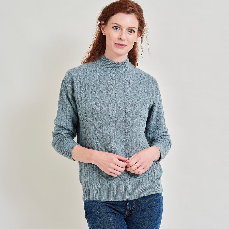 Woman wearing a light blue green cable knit cashmere sweater against a plain background