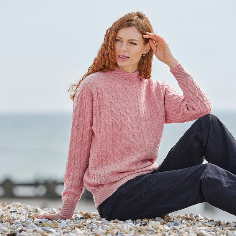 Woman wearing a pink cashmere sweater sitting on a pebbly beach.