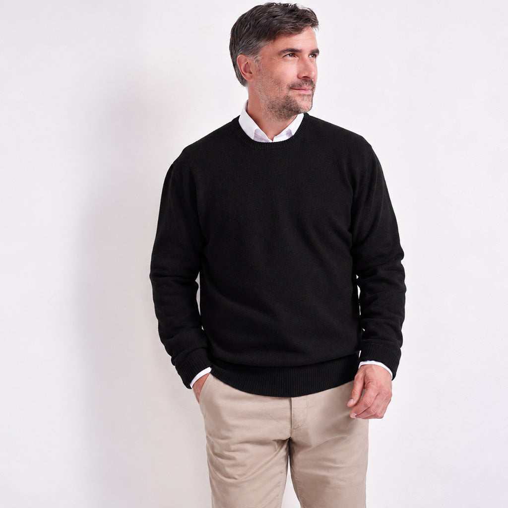 A man wearing a black crewneck sweater over a white collared shirt and beige trousers, standing against a plain white studio background.