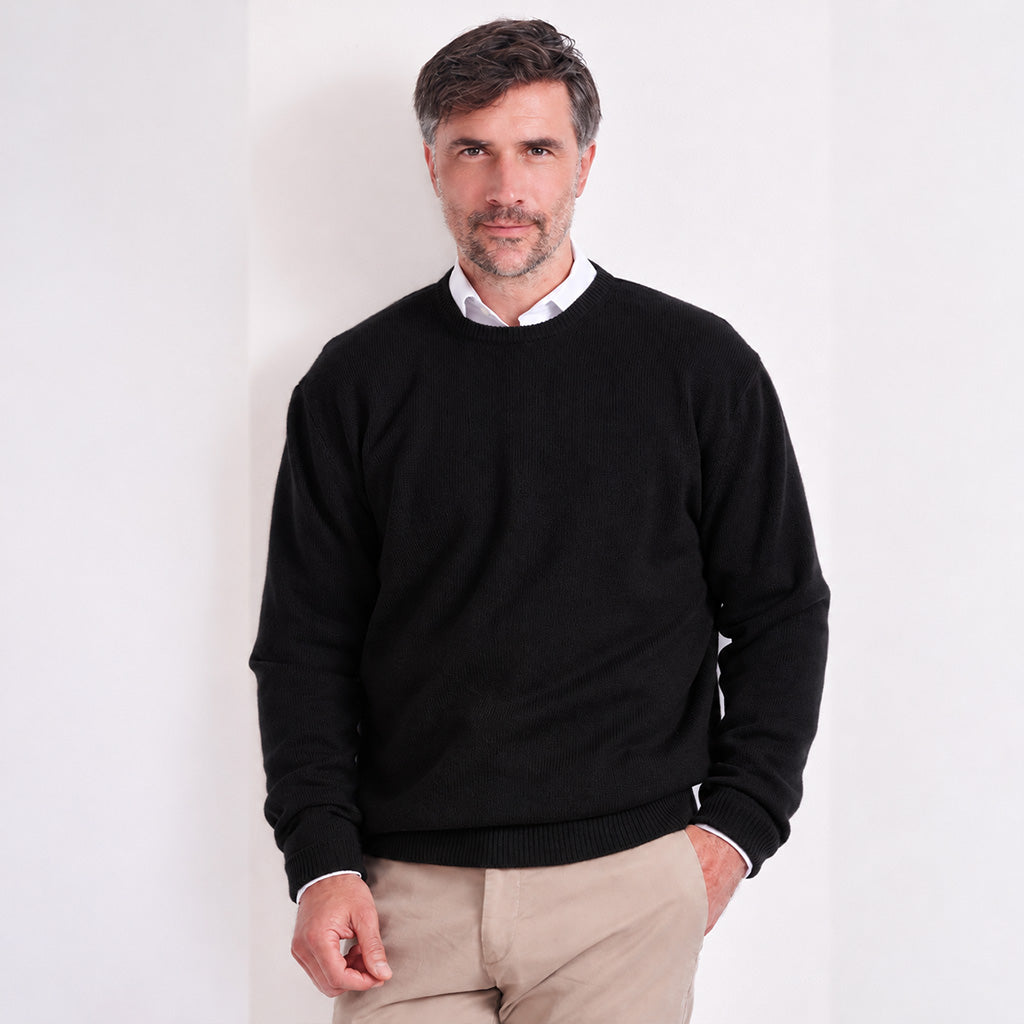 A man wearing a black crewneck sweater over a white collared shirt and beige trousers, standing against a plain white wall.