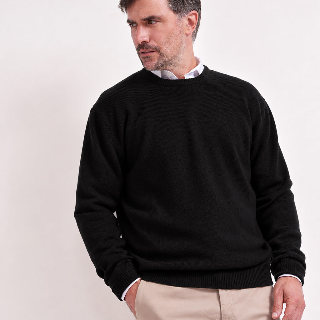 A man wearing a black crewneck sweater over a white collared shirt and beige trousers, photographed against a plain light background.