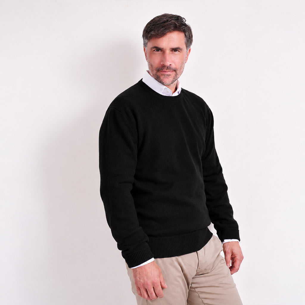 A man wearing a black crewneck sweater over a white collared shirt and beige trousers, standing against a plain white studio background.