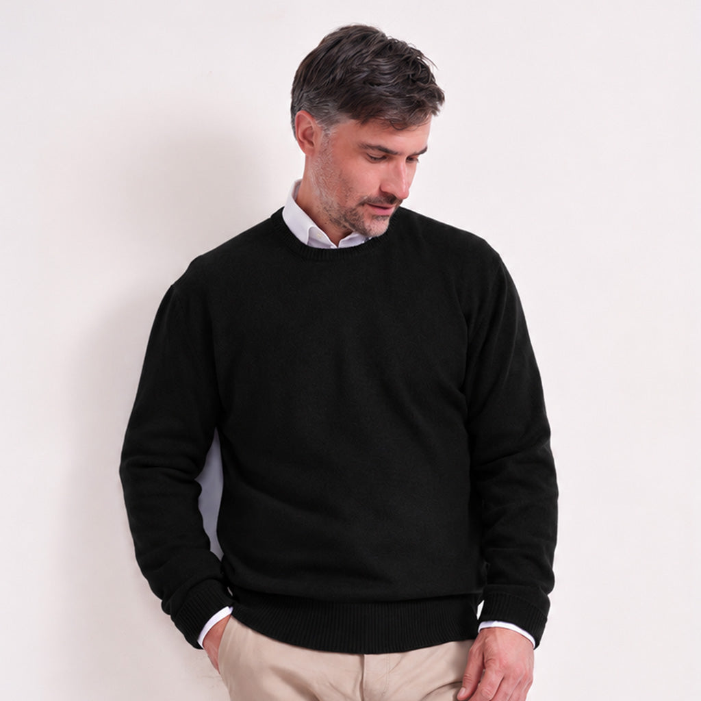 A man wearing a black crewneck sweater over a white collared shirt and beige trousers, standing against a plain off-white wall.