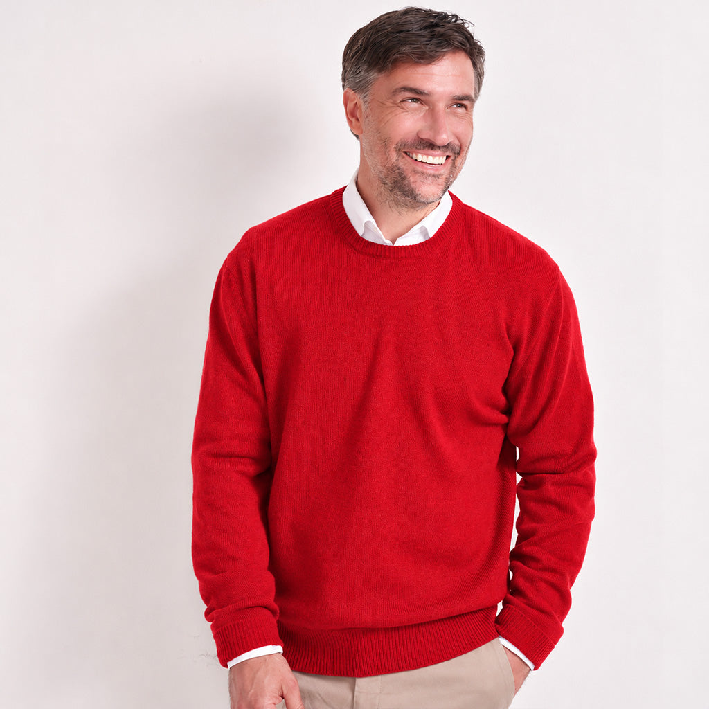 A smiling man wearing a bright red crewneck sweater over a white collared shirt and beige trousers, standing against a plain light gray studio background.