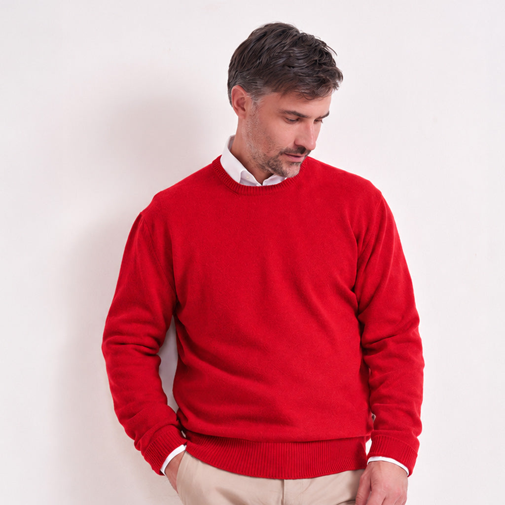A man wearing a bright red crewneck sweater over a white collared shirt and beige trousers, standing against a plain off-white wall.