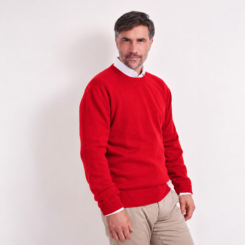 A smiling man wearing a bright red crewneck sweater over a white collared shirt and beige trousers, standing against a plain light gray studio background.