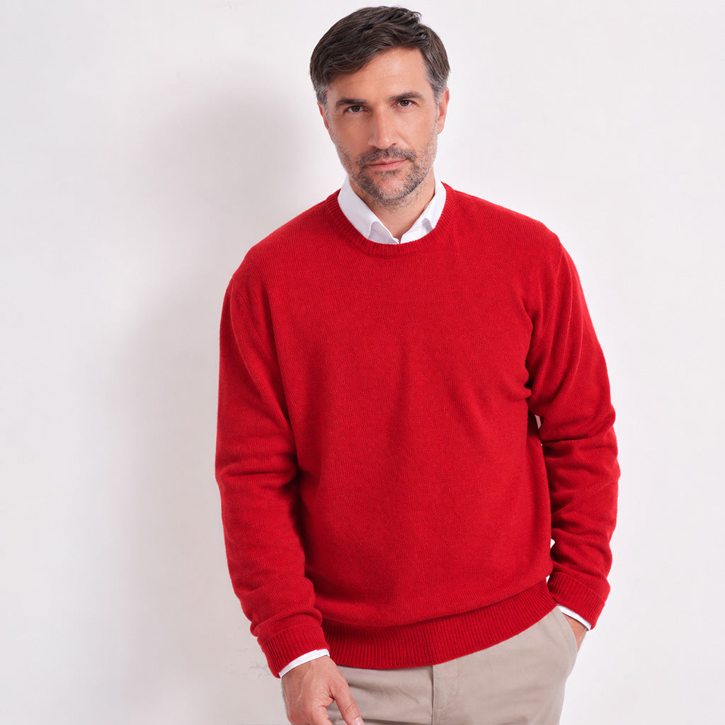 A smiling man wearing a bright red crewneck sweater over a white collared shirt and beige trousers, standing against a plain light gray studio background.
