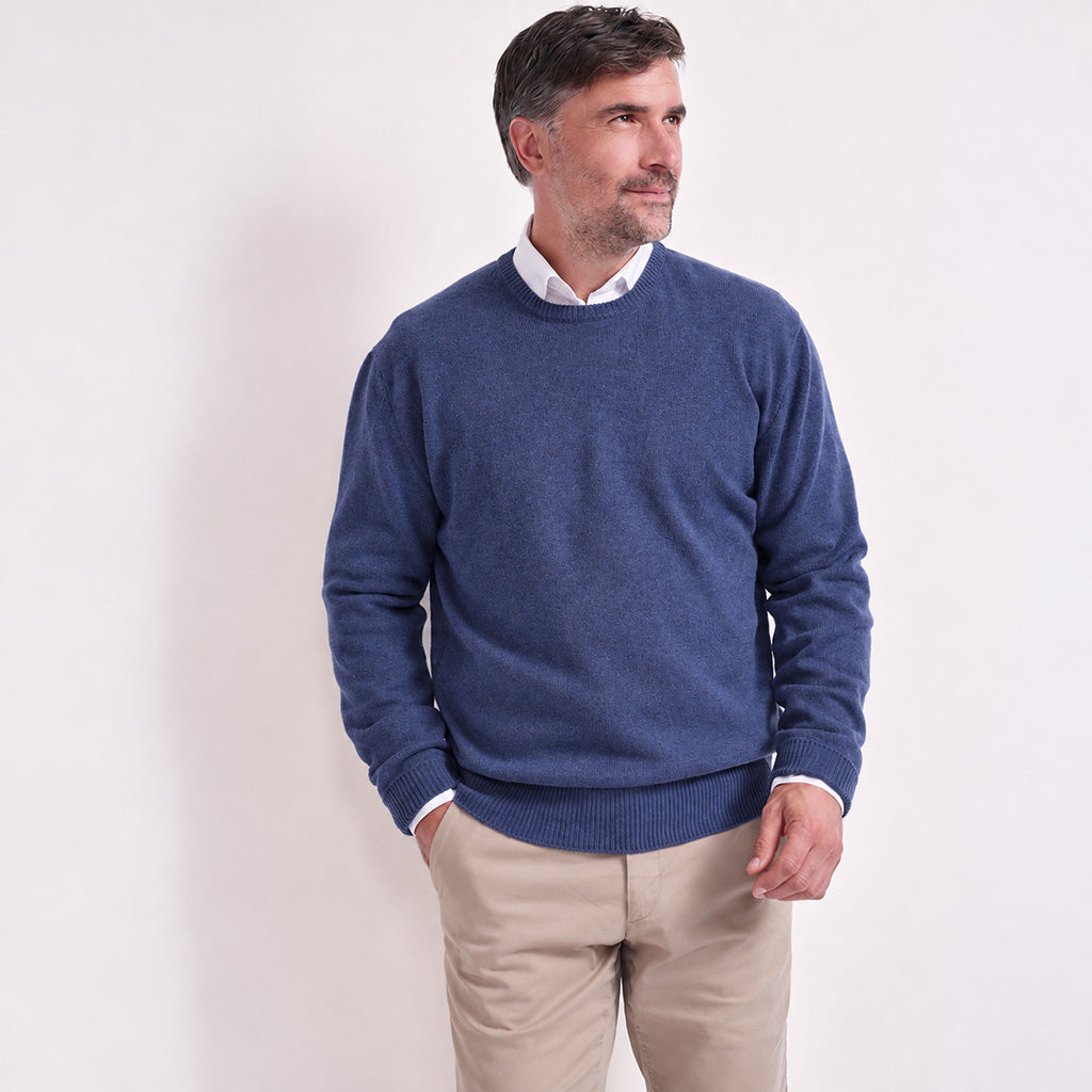 A man wearing a denim blue crewneck sweater over a white collared shirt and beige trousers, standing against a plain white studio background.