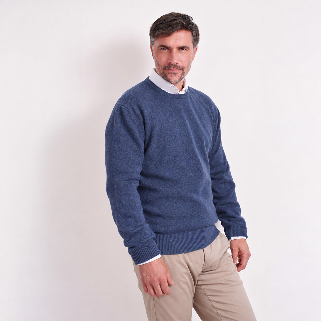 A man wearing a denim blue crewneck sweater over a white collared shirt and beige trousers, standing against a plain white studio background.