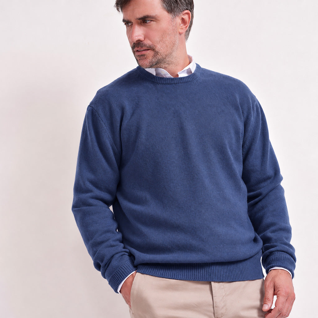 A man wearing a denim blue crewneck sweater over a white collared shirt and beige trousers, standing against a plain white studio background.