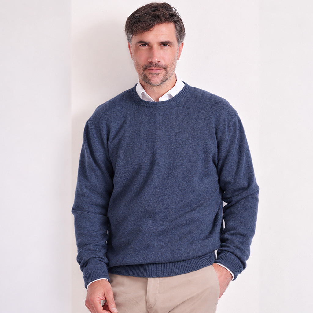 A man wearing a navy blue crewneck sweater over a white collared shirt and beige trousers, standing against a plain white wall.
