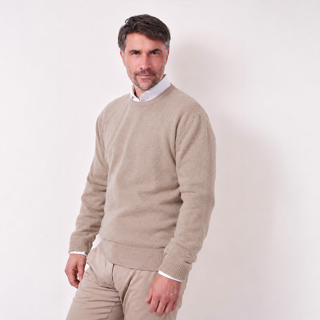 A man wearing a light beige knit crewneck sweater over a white collared shirt and beige trousers, standing against a plain off-white studio background.