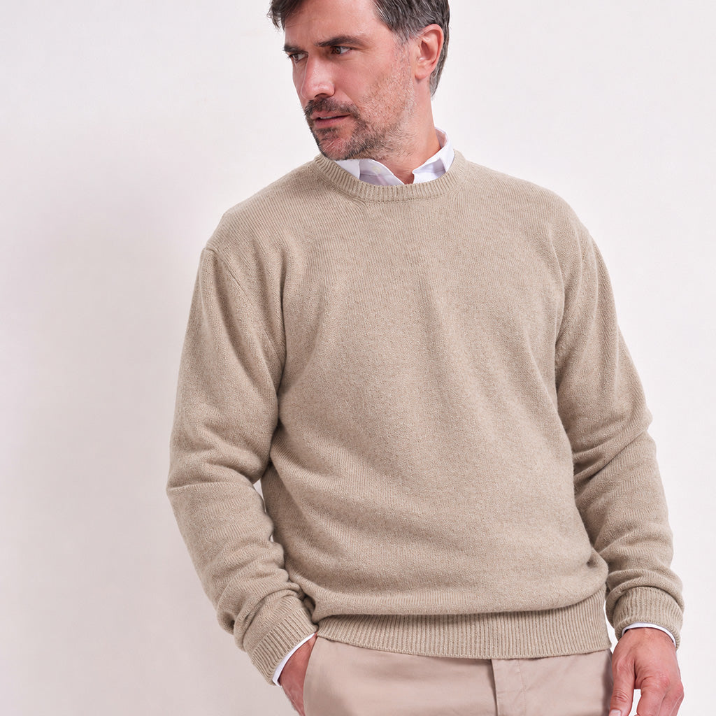 A man wearing a light beige crew-neck knit sweater over a white collared shirt, paired with beige trousers, photographed against a plain light background.