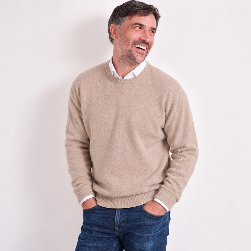 A smiling man wearing a light beige crewneck sweater over a white collared shirt and blue jeans, standing against a plain white background.