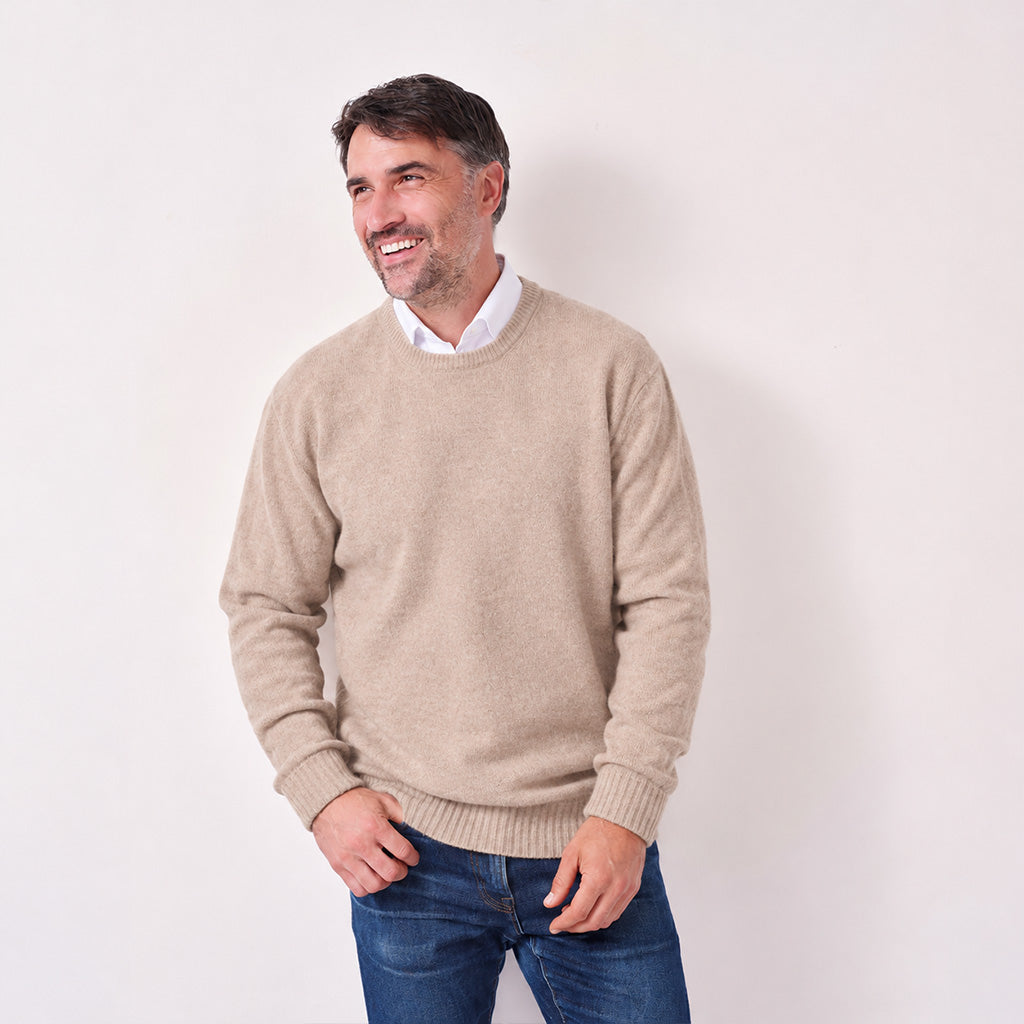 A smiling man wearing a light beige crewneck sweater over a white collared shirt and blue jeans, standing against a plain off-white wall.