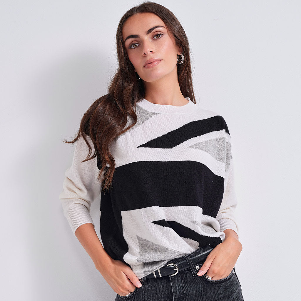A woman wearing a knit crewneck sweater with a modern abstract geometric pattern in black, off-white, and light gray, paired with dark jeans.