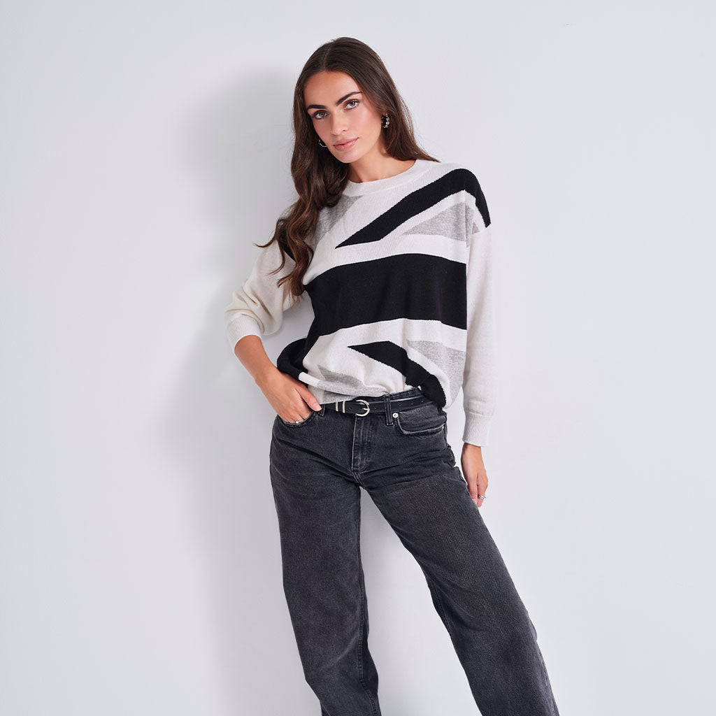 A woman wearing a knit crewneck sweater with a bold black, white, and gray geometric stripe pattern, posed against a light gray studio background.