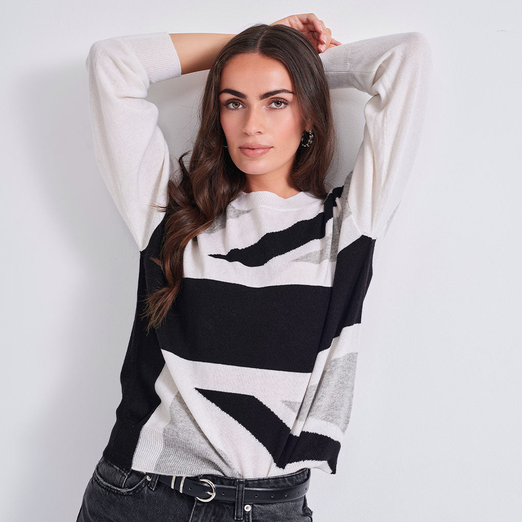 A woman wearing a knit crewneck sweater with a bold black, white, and gray geometric stripe pattern, posed against a light gray studio background.