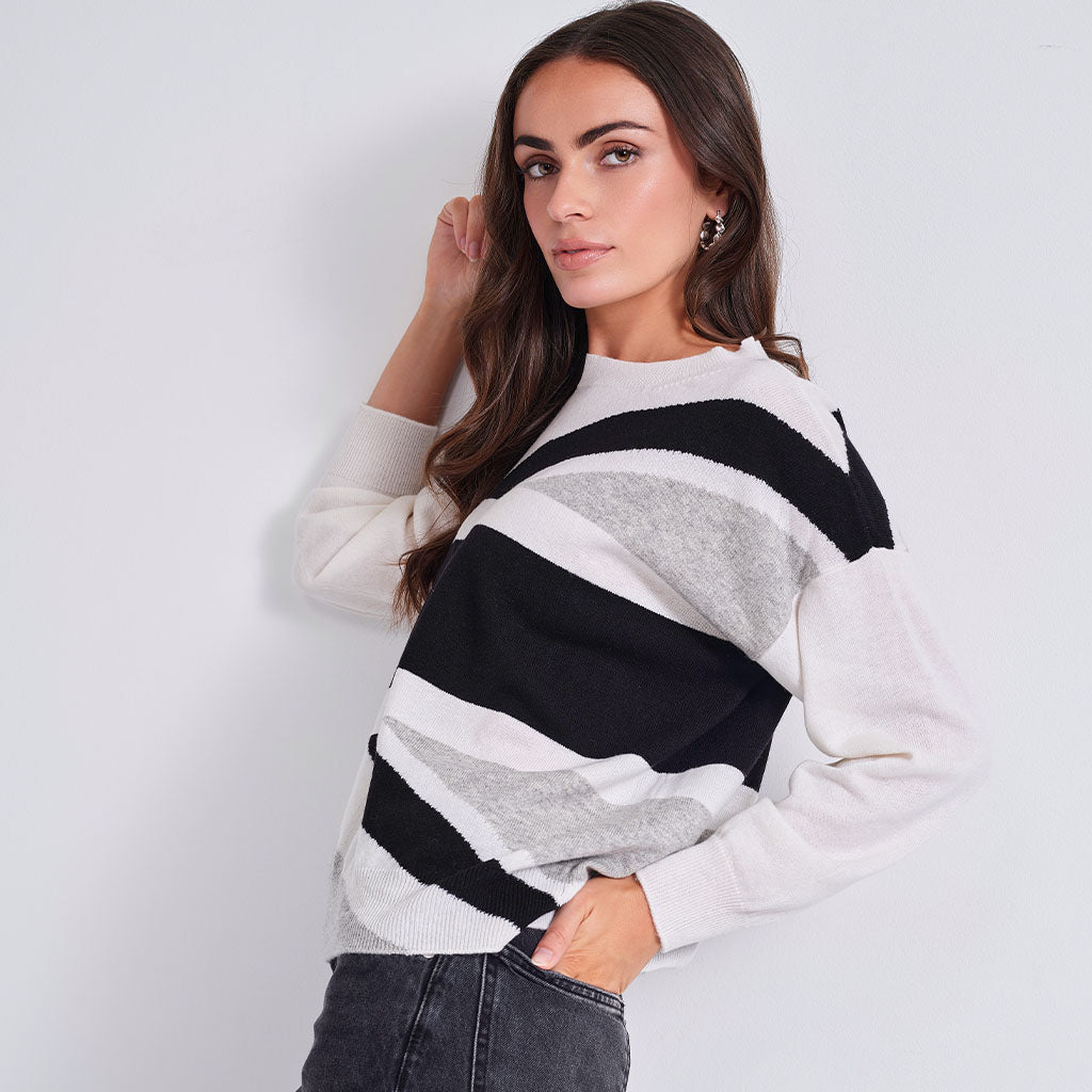 A woman wearing a long-sleeve knit sweater with bold horizontal stripes in black, white, and light gray, paired with dark gray jeans, posing against a light gray wall.