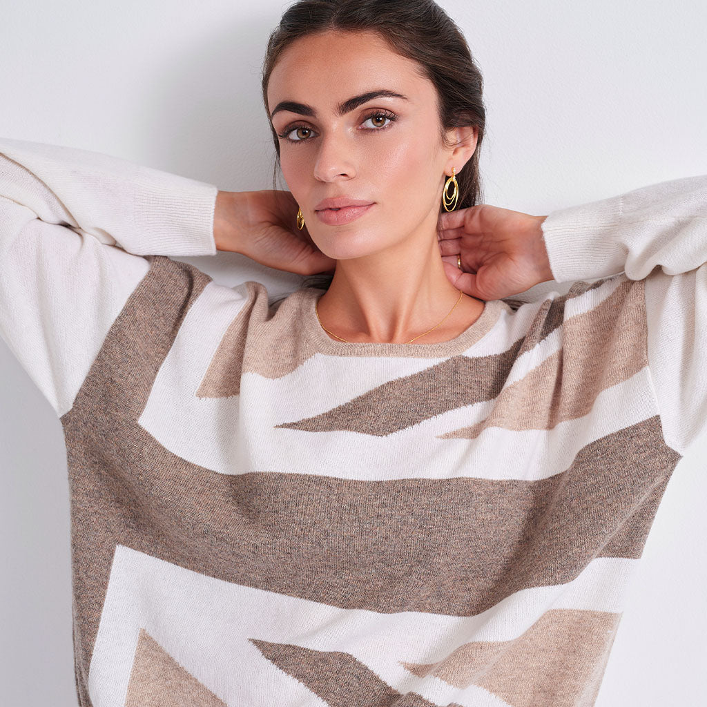 A woman wearing a knit crewneck sweater with a geometric abstract pattern in beige, taupe, and off-white, styled with gold hoop earrings against a plain light gray background.