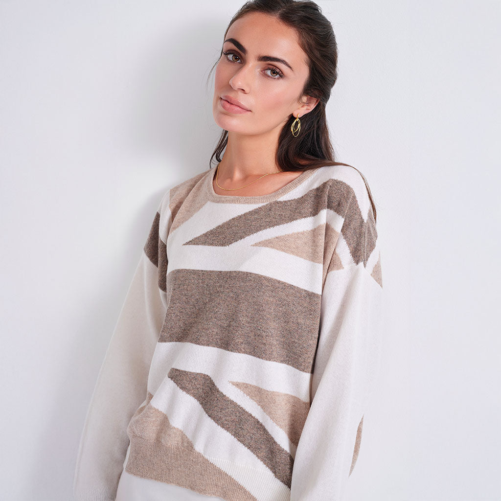A woman wearing a loose-fit knit sweater with wide sleeves and an abstract geometric stripe pattern in beige, taupe, and off-white, posed against a plain light gray wall.
