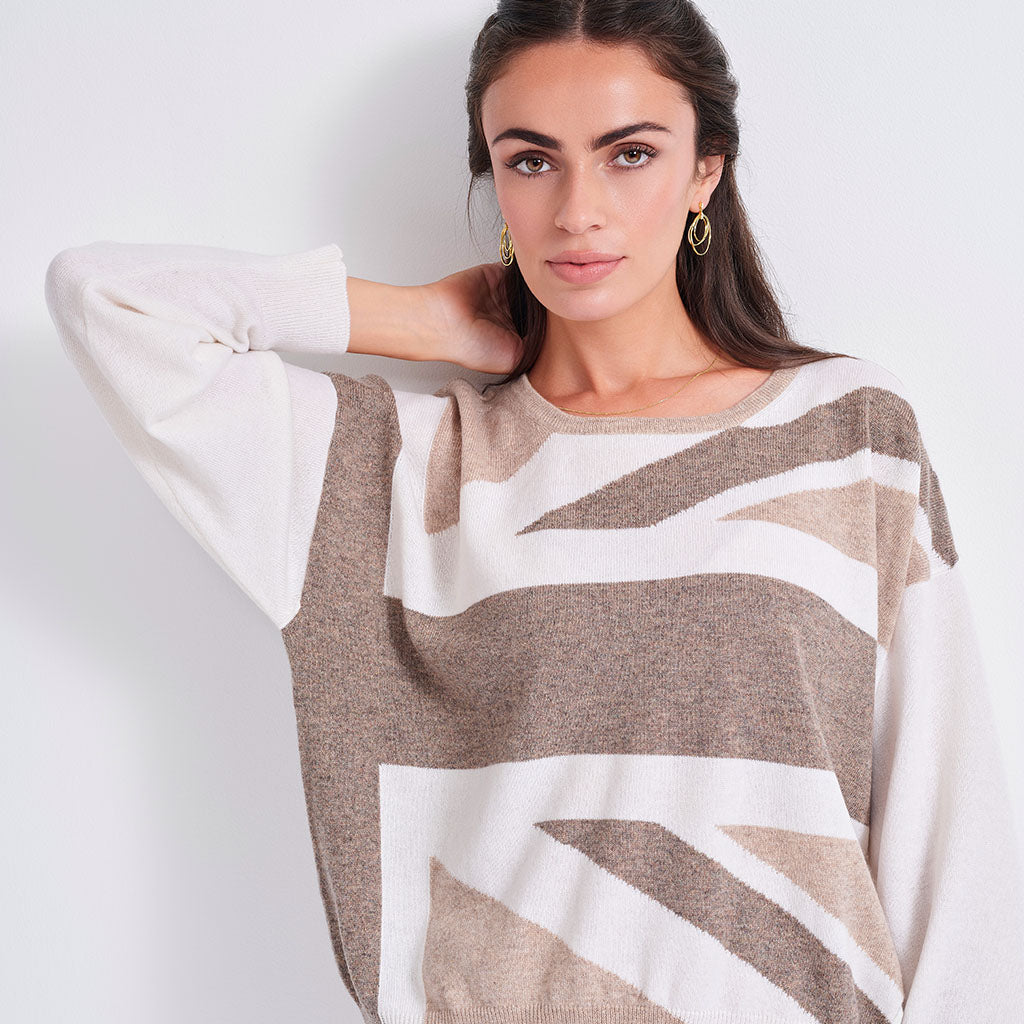 A woman wearing a knit sweater with a geometric abstract pattern in taupe brown and off-white, posed against a light gray studio background.