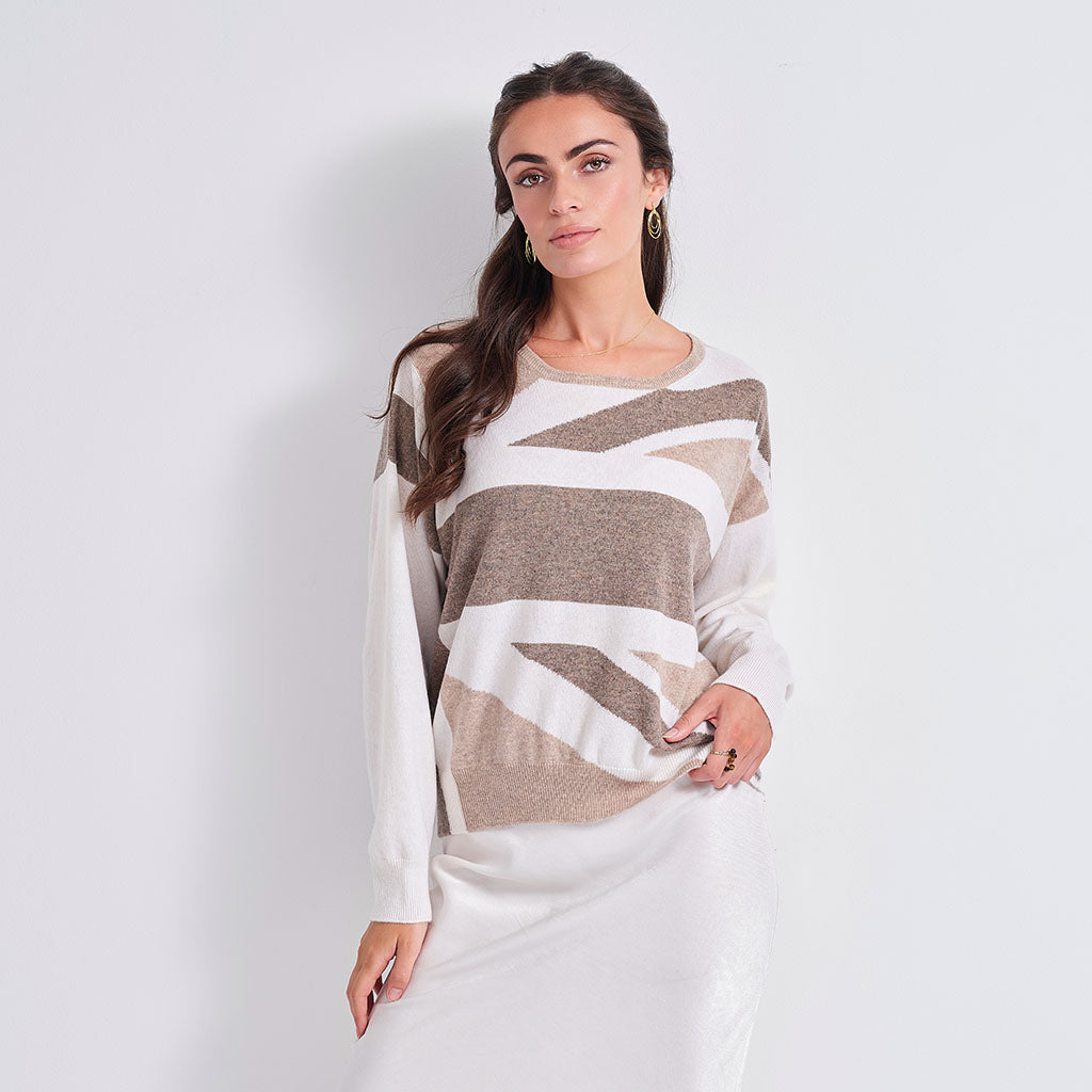 A woman modeling a long-sleeve knit sweater with a beige and white abstract geometric pattern, paired with a white skirt, against a plain light gray background.