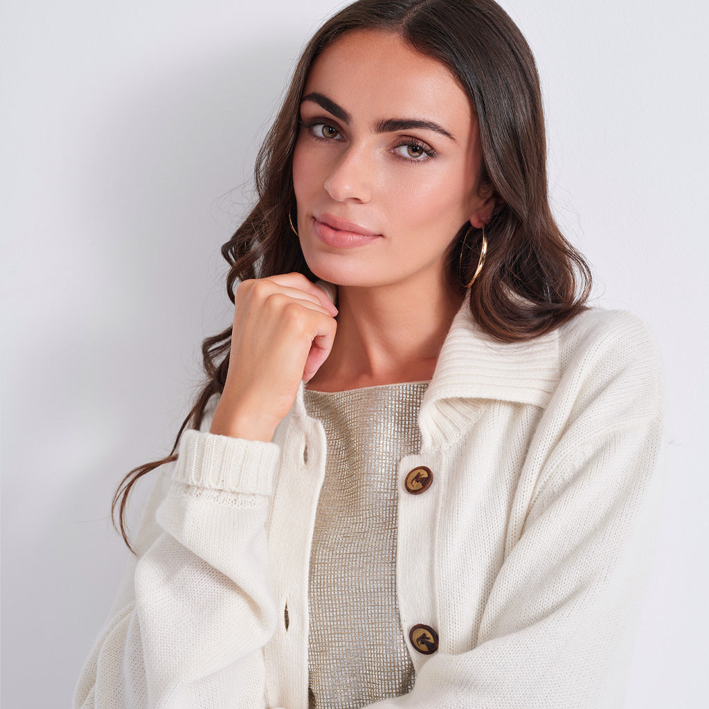 close up of a woman modeling an oversized cream-colored knit cardigan with brown buttons over a light beige top, set against a plain light gray studio background.