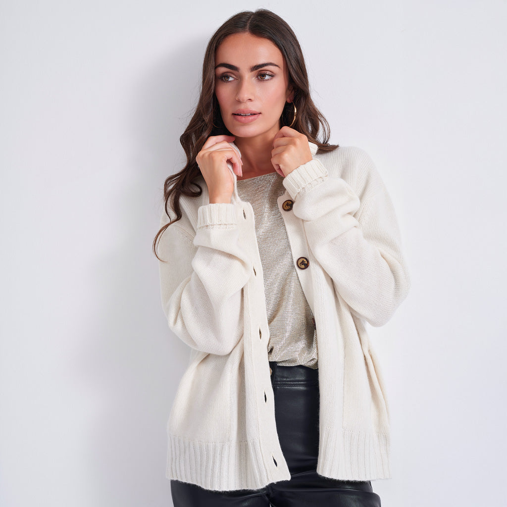 A woman modeling an oversized cream-colored knit cardigan with brown buttons over a light beige top and black pants, set against a plain light gray studio background.