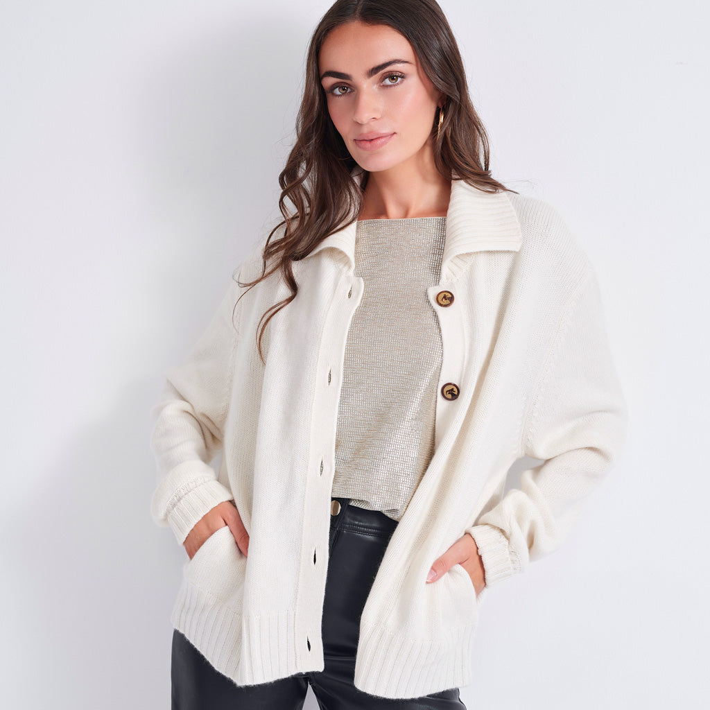 A woman modeling an oversized cream-colored knit cardigan with brown buttons over a light beige top and black pants, set against a plain light gray studio background.
