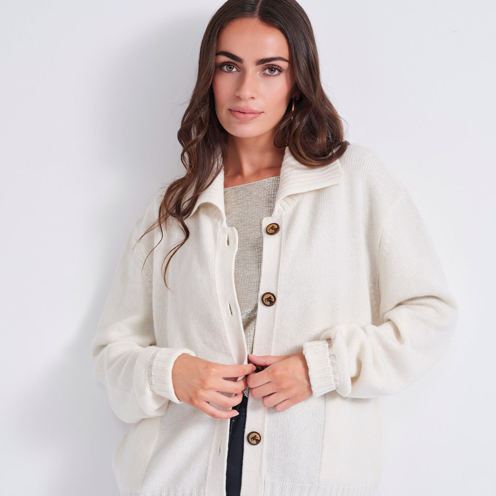 A woman wearing a cream-colored knit cardigan with brown buttons over a gray top, photographed against a plain light gray studio background.