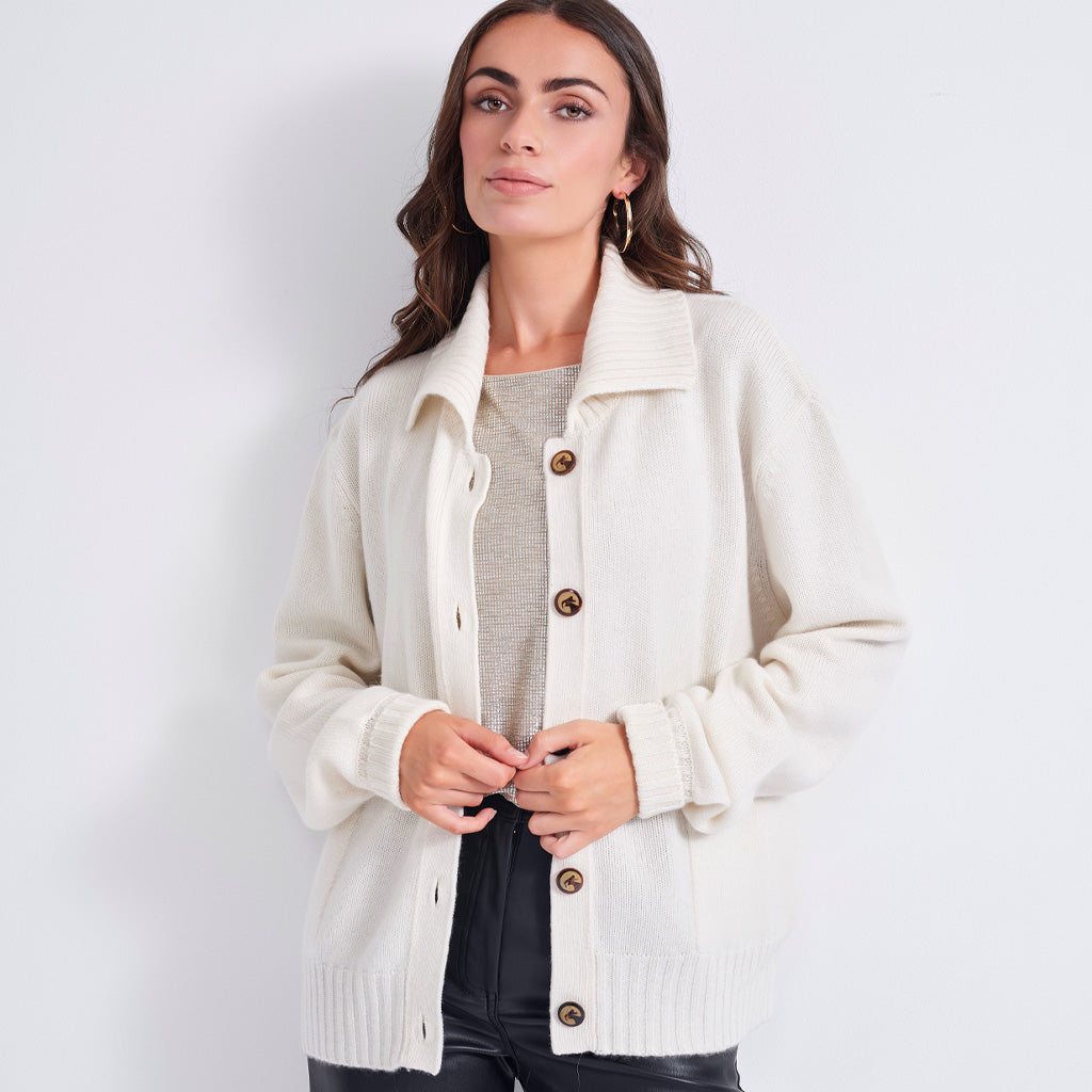 A woman wearing a cream-colored knit cardigan with brown buttons over a gray top and black pants, standing against a plain light gray background.
