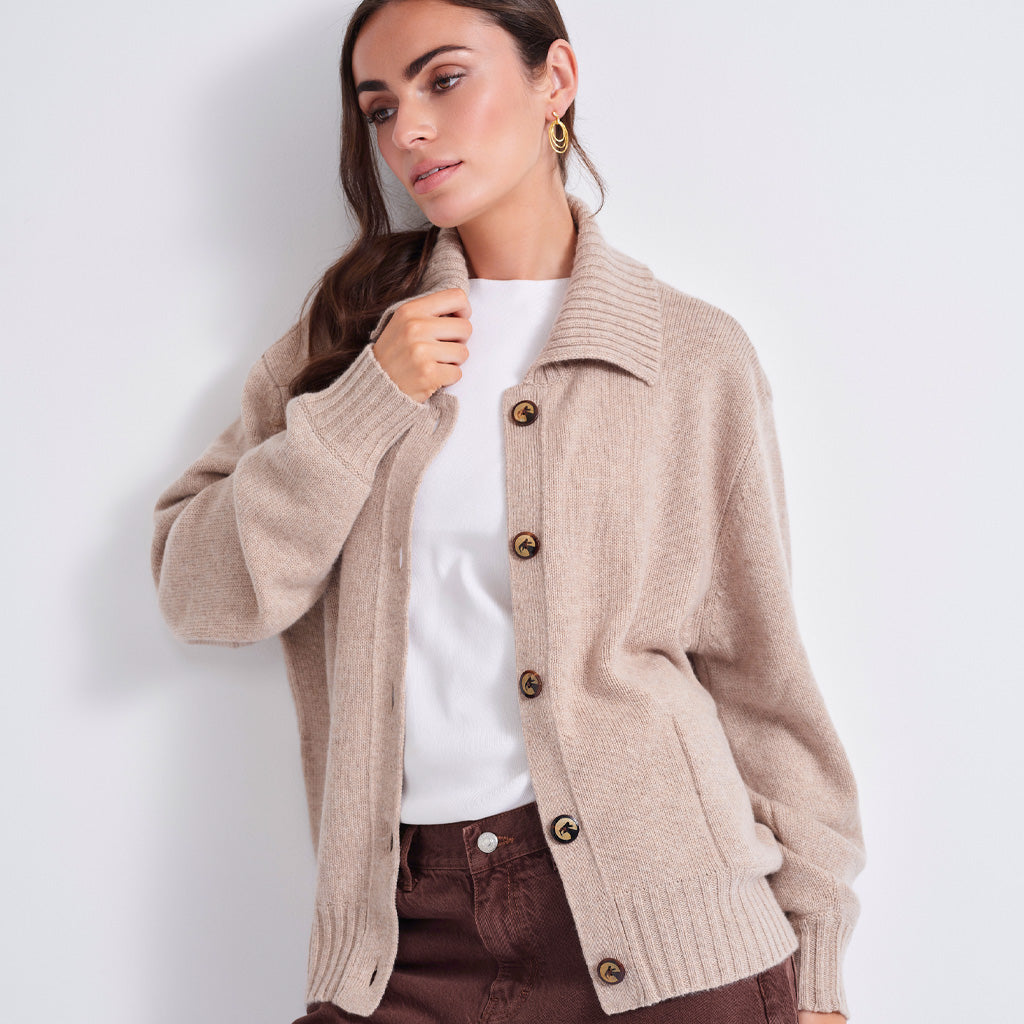 A woman wearing a beige knit button-front cardigan over a white top and brown pants, photographed against a plain light gray studio background.