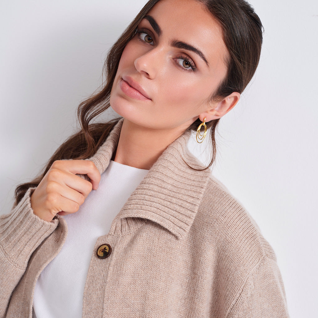 close up of a woman wearing a beige knit button-front cardigan over a white top and brown pants, photographed against a plain light gray studio background.