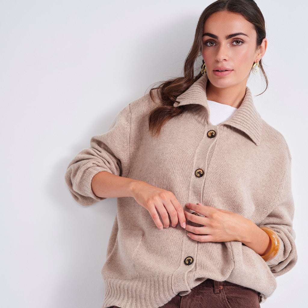 A woman models a light beige knit cardigan sweater with brown buttons, worn over a white top and brown pants, against a plain light gray background.