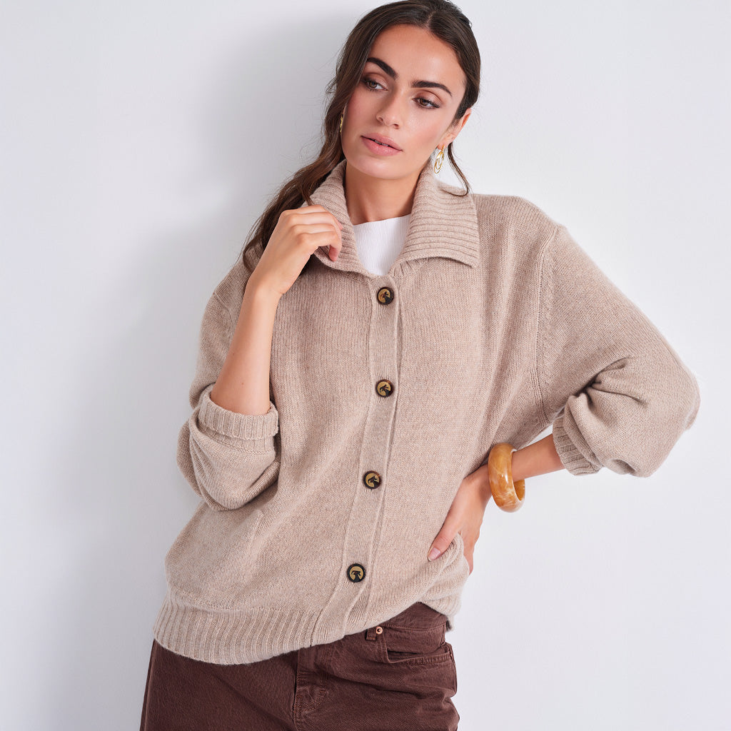 A woman models a light beige knit cardigan sweater with brown buttons, worn over a white top and brown pants, against a plain light gray background.