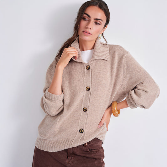 A woman models a light beige knit cardigan sweater with brown buttons, worn over a white top and brown pants, against a plain light gray background.