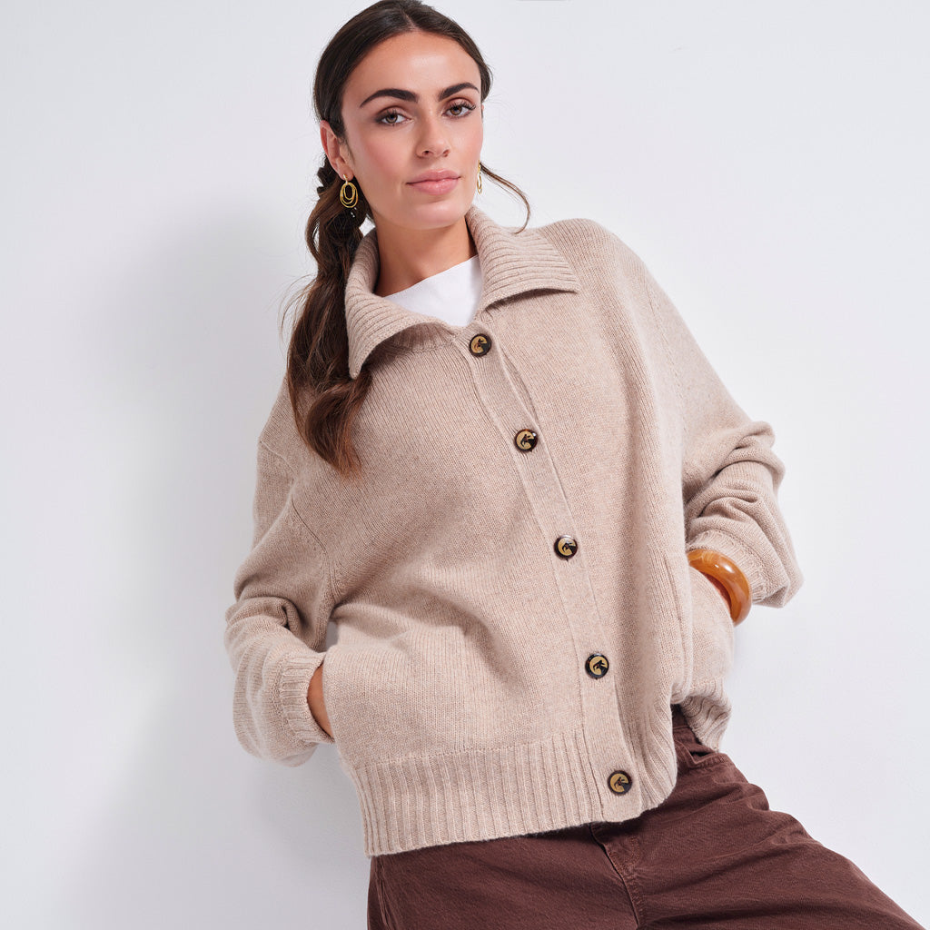 A woman models a light beige knit cardigan sweater with brown buttons, worn over a white top and brown pants, against a plain light gray background.