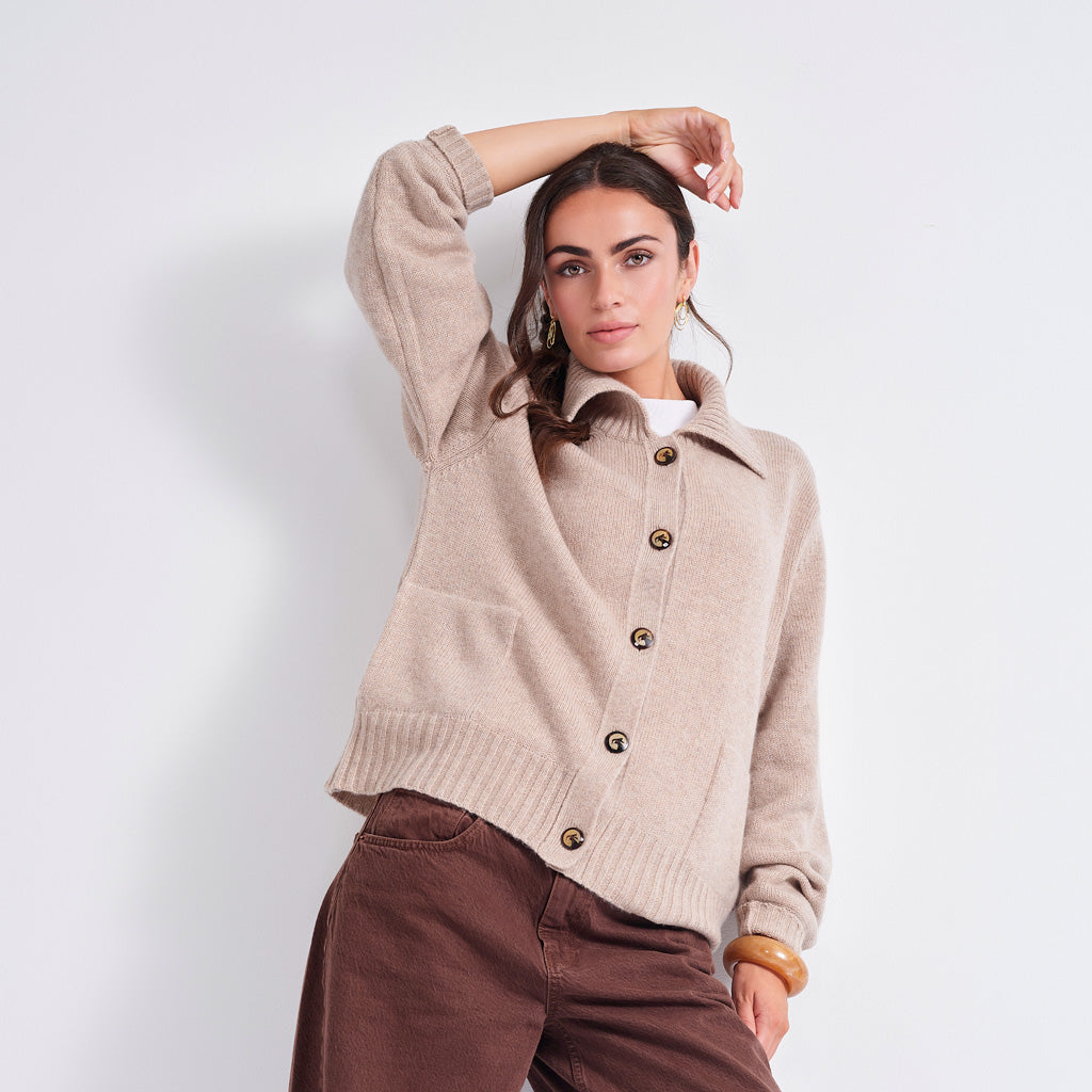 A woman models a beige knit button-front cardigan with a collar and brown buttons, paired with brown pants, against a plain light gray studio background.